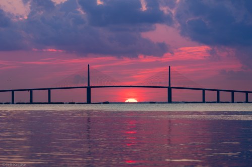 A vibrant sunset casts pink and orange hues behind a silhouetted bridge over calm waters.