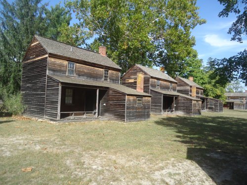 Four rustic wooden houses with porches, surrounded by grass and trees under a clear blue sky.