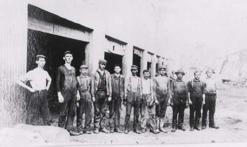 A historical black-and-white photo of a group of eleven male workers standing outside a building, with one woman on the left.