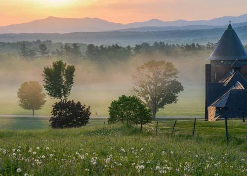 A misty landscape at sunrise, featuring trees, a distant figure walking, and a church with mountains in the background.