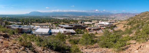 A panoramic view of a city with buildings and greenery, set against a backdrop of mountains and a clear blue sky.
