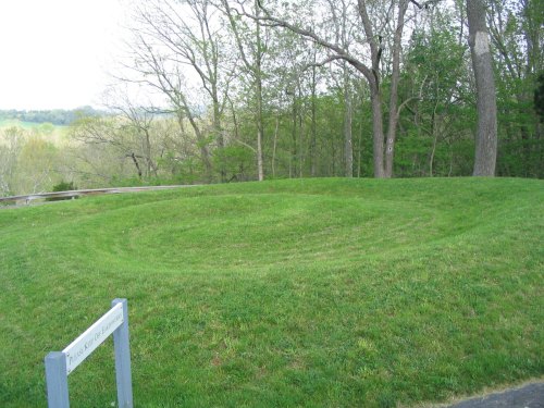 A grassy, circular earthwork surrounded by trees, with a sign nearby indicating its historical significance.