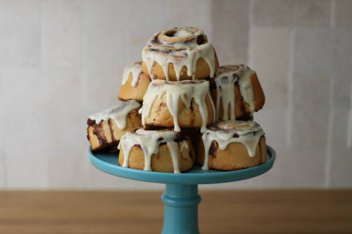 A stack of cinnamon rolls with cream cheese icing on a blue cake stand, set against a light tiled background.