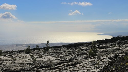 A rocky landscape with sparse vegetation, overlooking a shimmering ocean under a blue sky with scattered clouds.