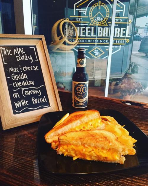A plate of grilled cheese with mac and cheese, gouda, and cheddar, alongside a bottle of beer and a menu sign.