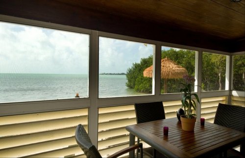 A cozy porch with a wooden table and chairs, overlooking a calm sea and lush greenery under a cloudy sky.