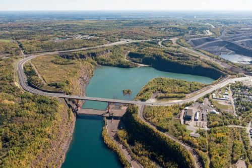 Aerial view of a winding road and bridge over a lake, surrounded by lush greenery and mining sites in the background.