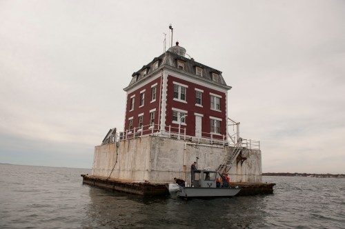 A red and white lighthouse on a concrete base in the water, with a small boat nearby and overcast skies.