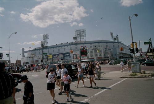 Crowd of people crossing the street near a large stadium on a sunny day, with cars and buildings in the background.
