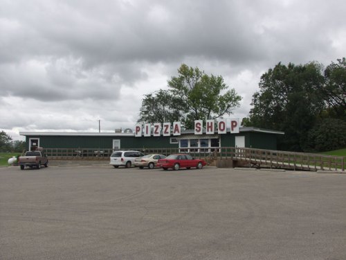 A green pizza shop building with large "PIZZA SHOP" sign, surrounded by a parking lot and cloudy sky.