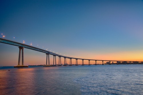 A long bridge spans over calm water at sunset, with lights twinkling against a gradient sky.