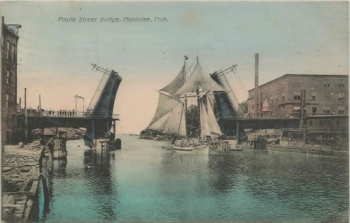 A vintage postcard of Maple Street Bridge in Manistee, Michigan, with a sailboat passing under the raised bridge.