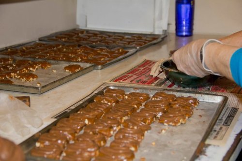 A person in gloves drizzles chocolate over rows of treats on trays in a kitchen setting.