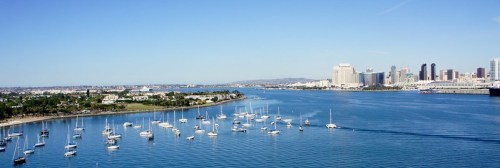 A panoramic view of a harbor filled with sailboats, with a city skyline in the background under a clear blue sky.