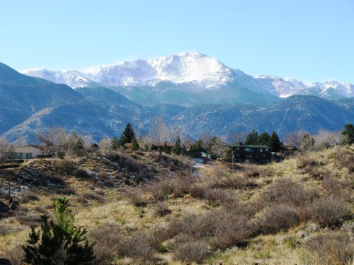 A scenic view of snow-capped mountains in the background, with a grassy foreground and scattered trees and houses.