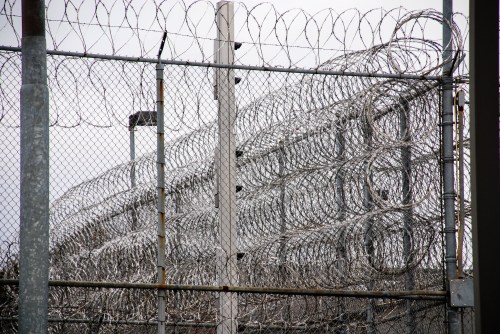 A close-up of a barbed wire fence surrounding a prison, with a cloudy sky in the background.