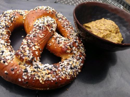 A large, salted pretzel topped with seeds, served with a small bowl of mustard on a dark plate.