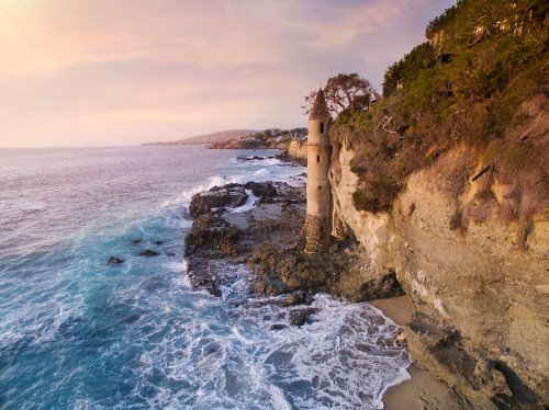 A coastal scene featuring a stone tower on a cliff, with waves crashing against the rocks and a colorful sunset sky.