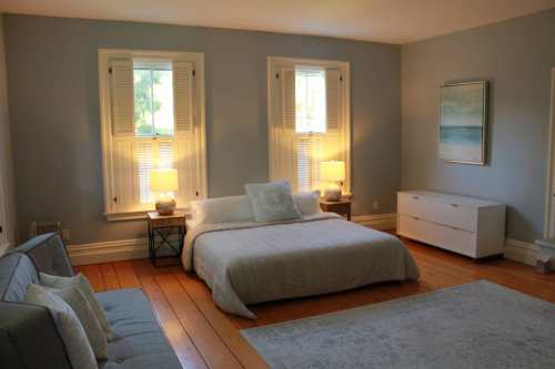 Bright, airy bedroom with a bed, two lamps, a dresser, and large windows, featuring light blue walls and wooden floors.