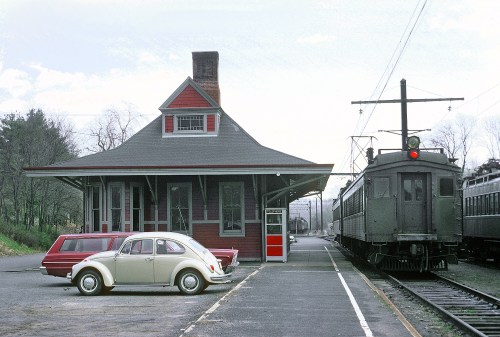 A vintage train station with a red and gray building, a classic car parked nearby, and a train approaching on the tracks.