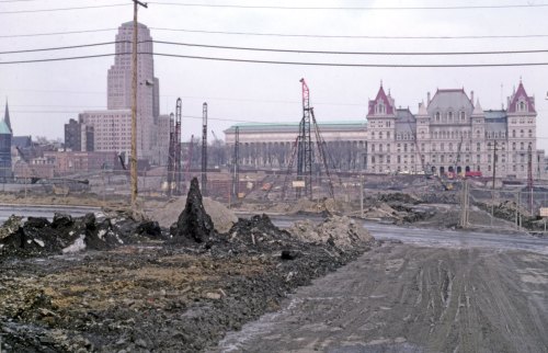 Construction site with heavy machinery, muddy ground, and buildings in the background, under a cloudy sky.