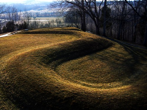 A grassy, undulating landscape with gentle hills and shadows, surrounded by trees and open fields in the background.