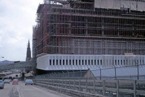 Construction site with scaffolding, large building structure, and cars on a nearby road under a cloudy sky.