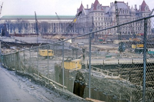 Construction site with cranes and machinery, surrounded by a chain-link fence, in a cityscape setting.