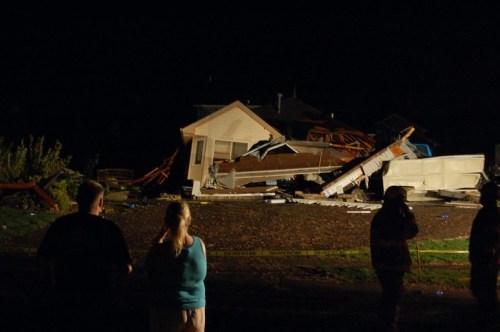 A damaged house at night, with debris scattered and onlookers observing the destruction.