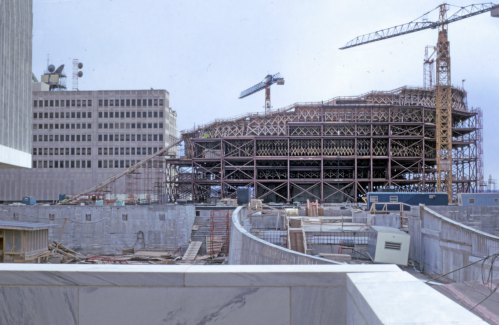 Construction site featuring cranes and scaffolding around a large, partially built structure, with surrounding buildings.