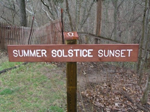 Wooden sign reading "Summer Solstice Sunset" in a wooded area with a path leading into the trees.