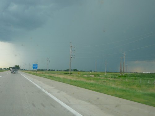 A highway scene with dark storm clouds looming, power lines along the roadside, and a hint of rain in the distance.