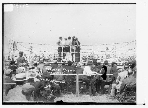 Historic boxing match with two fighters in the ring, surrounded by spectators in early 20th-century attire.