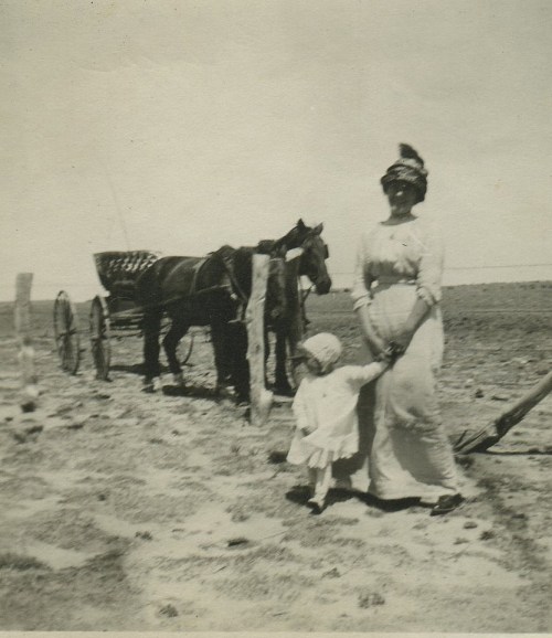 A woman in a long dress stands with a child by a horse-drawn cart in a barren landscape.