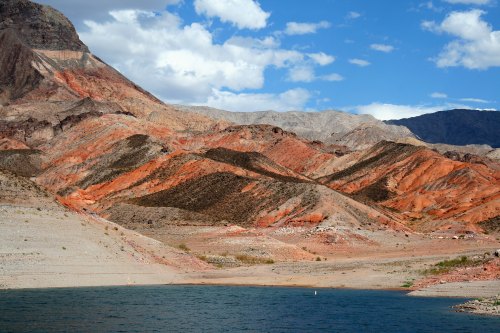 Vibrant red and orange mountain ridges rise above a calm blue lake under a partly cloudy sky.