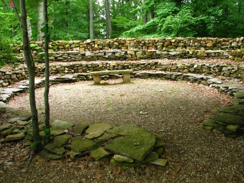 A circular stone seating area surrounded by trees, with a central stone bench and gravel ground.