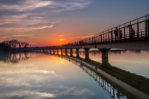 A sunset casts vibrant colors over a calm river, reflecting a bridge and silhouetted figures walking along it.