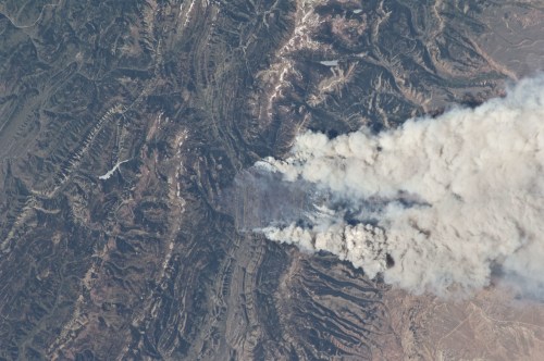 Aerial view of a mountainous landscape with thick smoke billowing from a wildfire, surrounded by rugged terrain.