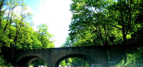 A stone bridge arches over a road, surrounded by lush green trees under a bright blue sky.