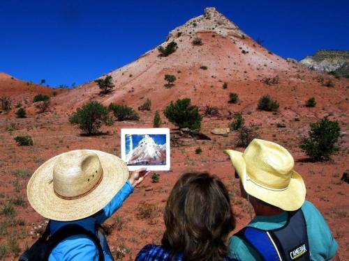 Two people in hats hold a photo of a mountain while standing in a red desert landscape with sparse vegetation.