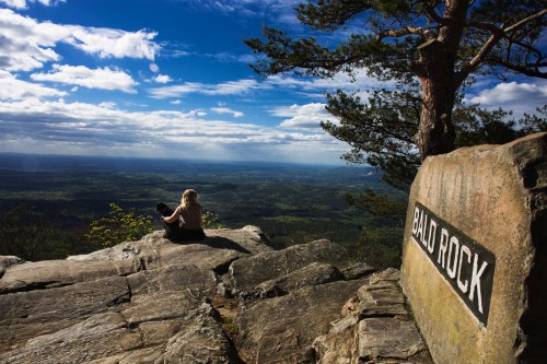 A person sits on a rocky ledge at Bald Rock, overlooking a vast landscape under a blue sky with scattered clouds.
