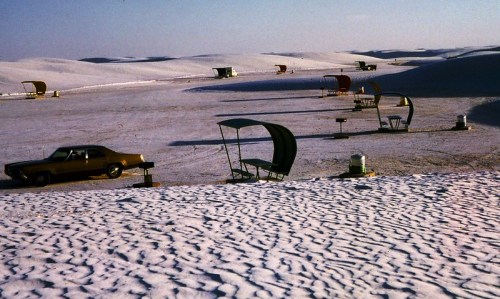 A vintage car parked on a snowy landscape with colorful chairs and tables scattered across the dunes.