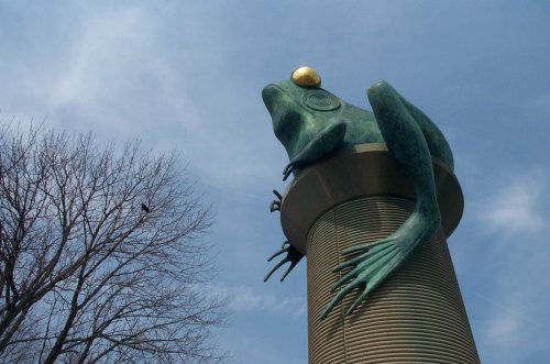 A large bronze frog sculpture perched atop a cylindrical pedestal against a blue sky.