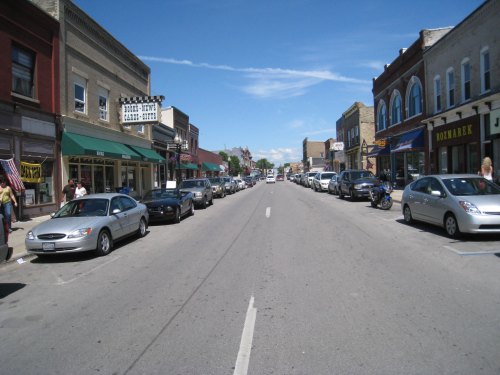 A wide street lined with shops and parked cars under a clear blue sky. People walk along the sidewalks.