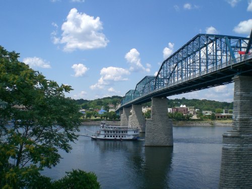A blue bridge spans a river with a steamboat below, surrounded by green trees and a clear blue sky with clouds.