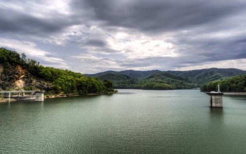 A serene lake surrounded by lush green mountains under a cloudy sky, with a dam structure on the left.