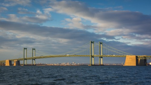 A suspension bridge spans a river under a blue sky with clouds, showcasing its green towers and cables.