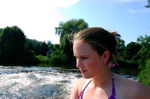 A young girl in a swimsuit gazes thoughtfully by a river with lush greenery and a blue sky in the background.