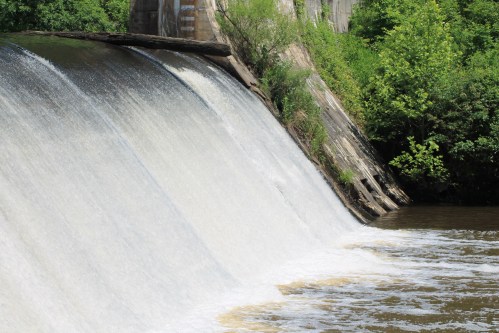 A cascading waterfall flows over a dam, surrounded by lush green trees and a calm river below.