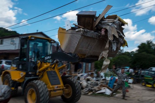 A loader lifts debris while a person gestures nearby, amidst a scene of cleanup after a disaster.